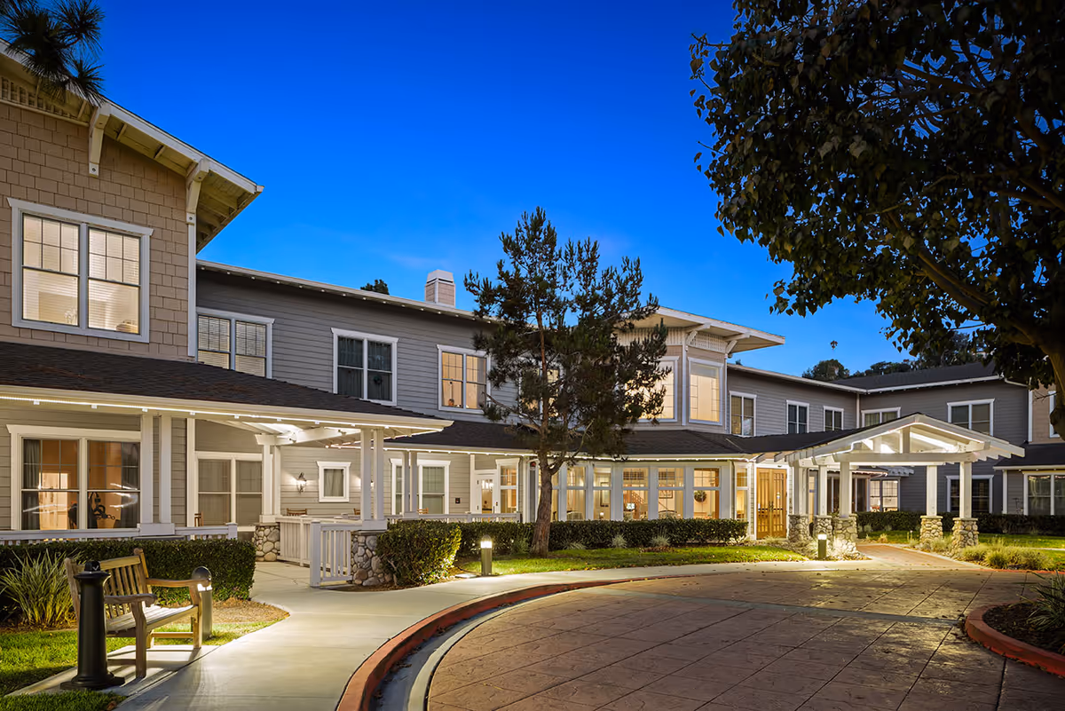 Front exterior of a two-story senior living facility at dusk showing the lit entrance, covered porte-cochere, walkway, and landscaping.
