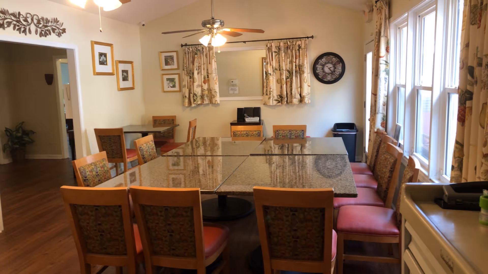 A dining room with a large rectangular table made of four smaller tables pushed together, surrounded by wooden chairs with patterned cushions and red seats. The room has hardwood floors, floral curtains on the windows, a wall clock, framed pictures, and a ceiling fan with lights. There is a trash bin in the corner and a small counter with a printer and a green container on the right side.