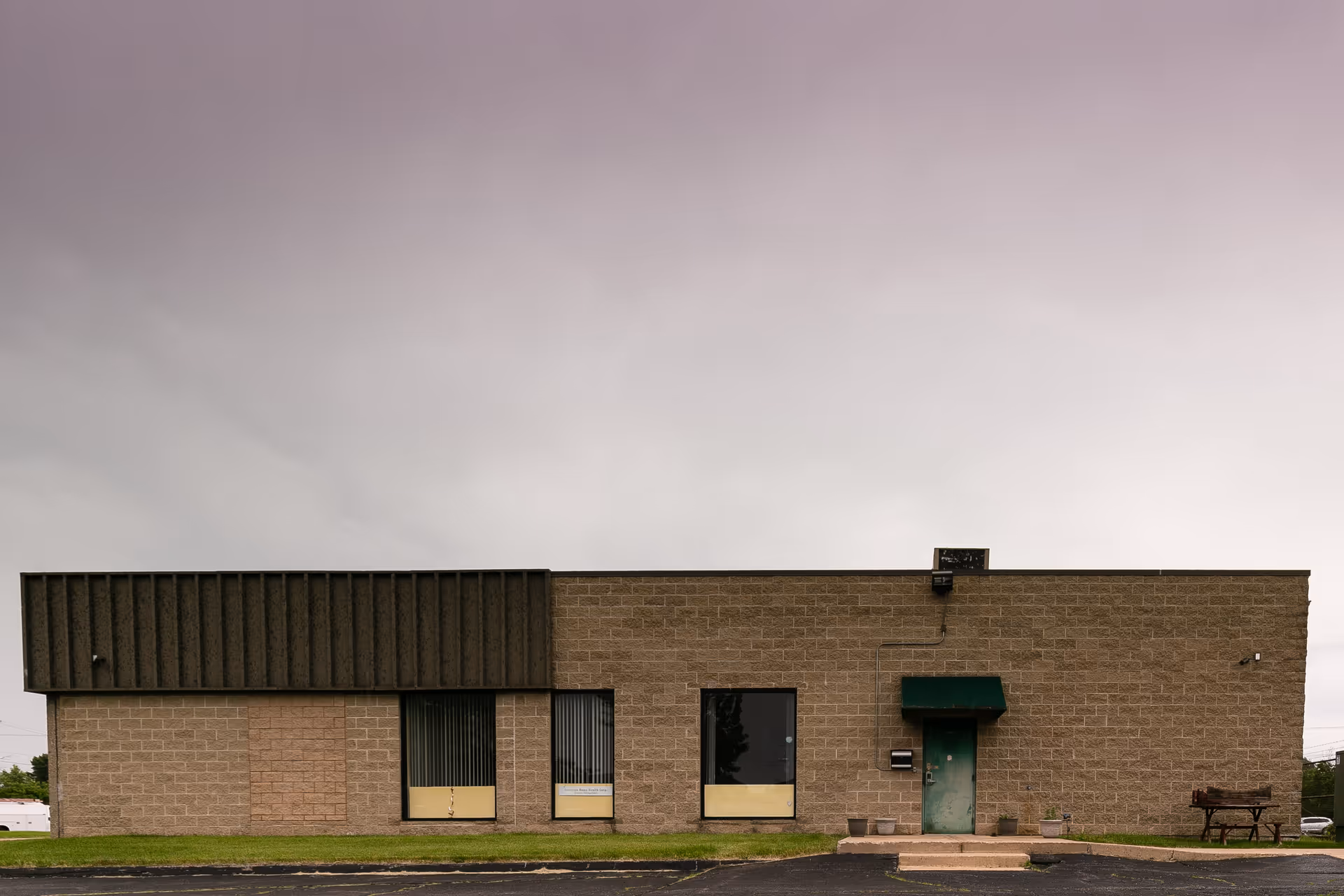 A single-story brick facility with green door and bench in front under a cloudy sky