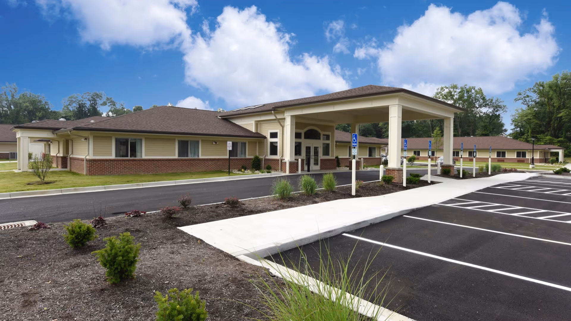 Exterior view of Liberty Nursing Center of Colerain showing a single-story building with beige siding and brick accents, a covered entrance with columns, a paved parking lot with marked handicap spaces, landscaped areas with small shrubs, and a partly cloudy blue sky.