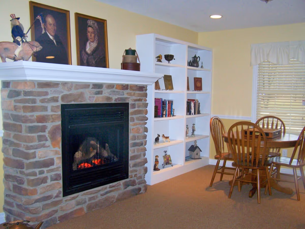 Cozy room with a stone fireplace featuring a black metal insert and a white mantel. Above the mantel are two framed portraits and a decorative figure of a person on a horse. To the right of the fireplace is a white built-in bookshelf filled with books and various decorative items. A wooden dining table with four matching chairs is positioned near a window with white blinds and a valance. The walls are painted light yellow and the floor is carpeted.