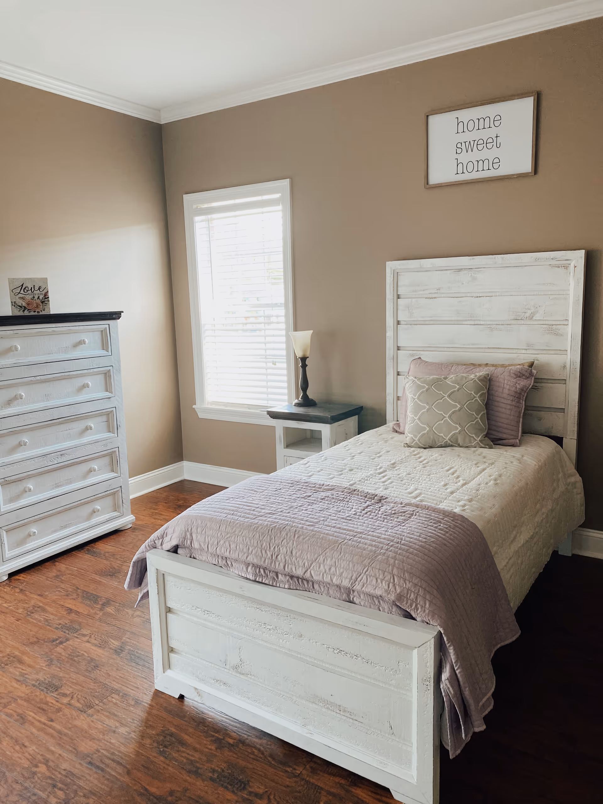 Sunlit single bedroom with a white wooden bed, matching dresser and nightstand, a lamp, and a 'home sweet home' wall sign.