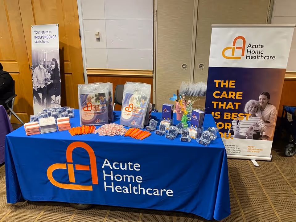 Promotional booth for Acute Home Healthcare with brochures and gifts on a blue table.