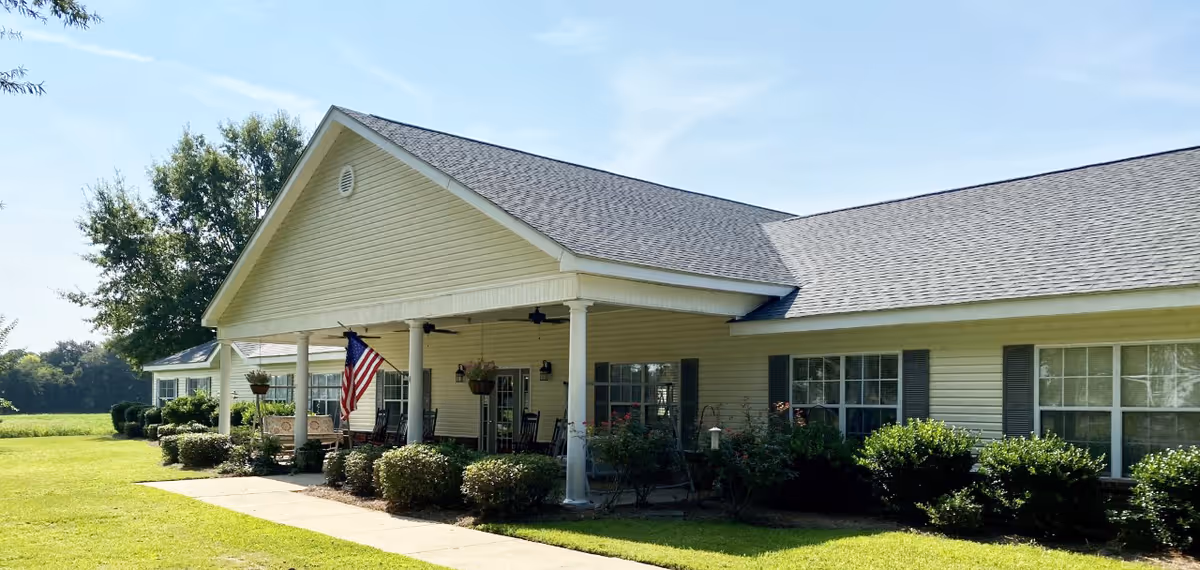 Exterior view of a single-story building with a covered porch, beige siding, and a gray shingled roof. The porch has several rocking chairs, hanging plants, and an American flag. The building is surrounded by neatly trimmed bushes and a well-maintained lawn under a clear blue sky.