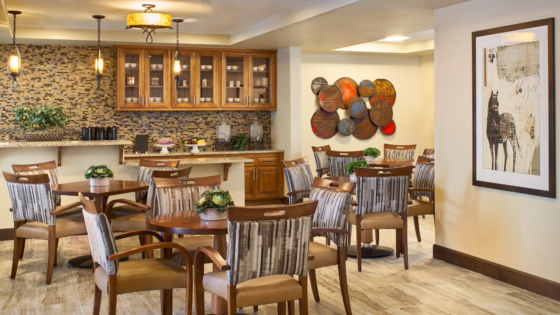 Bright dining area in a senior living facility with wooden tables, striped chairs, and a kitchenette.