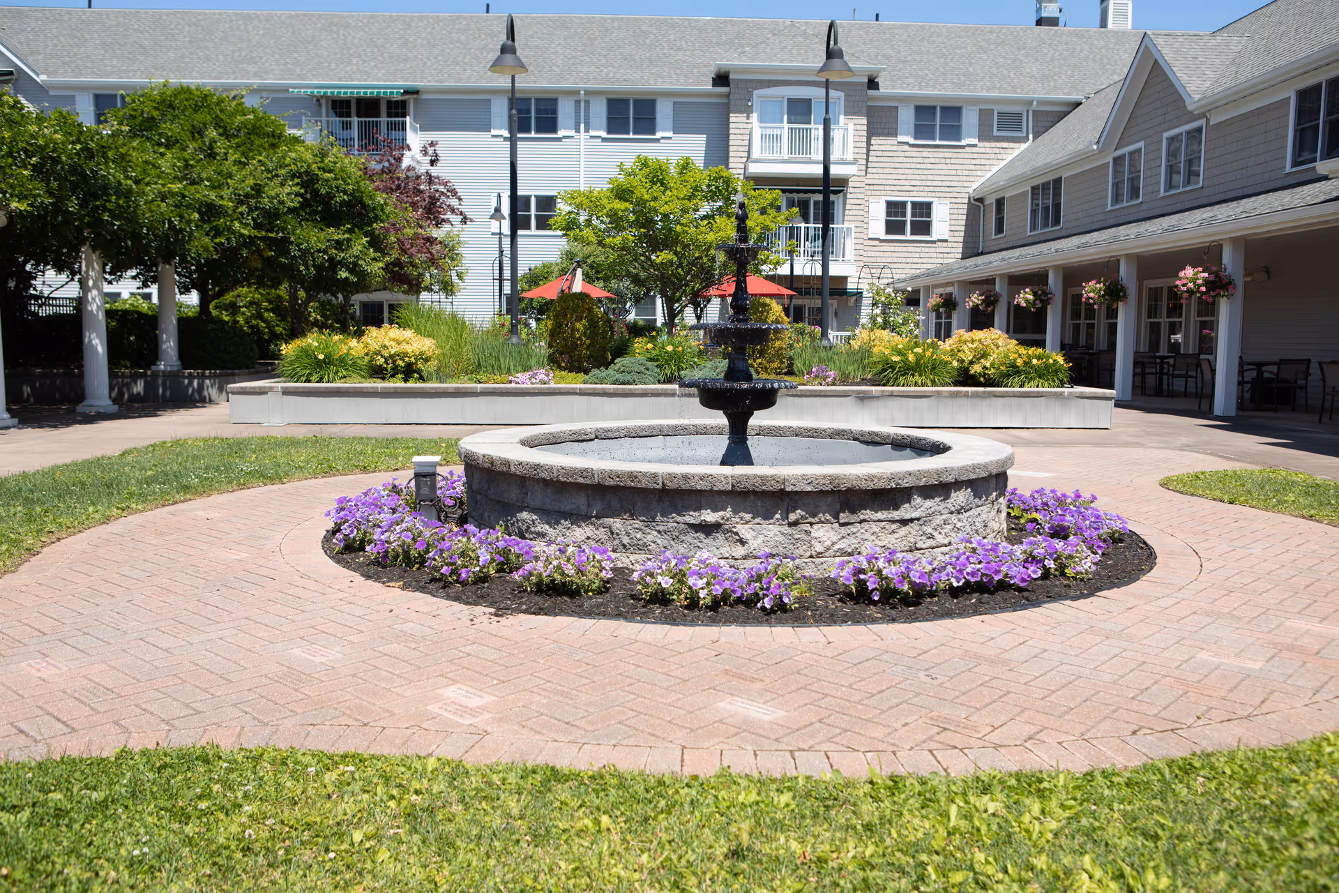 Circular stone fountain in brick courtyard, surrounded by flowers and senior living buildings.