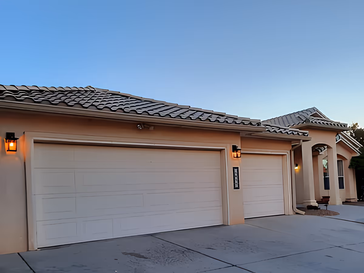 Exterior view of a residential building with a tiled roof and three closed garage doors. The building has beige walls and outdoor wall-mounted lights beside each garage door, with a clear blue sky in the background.
