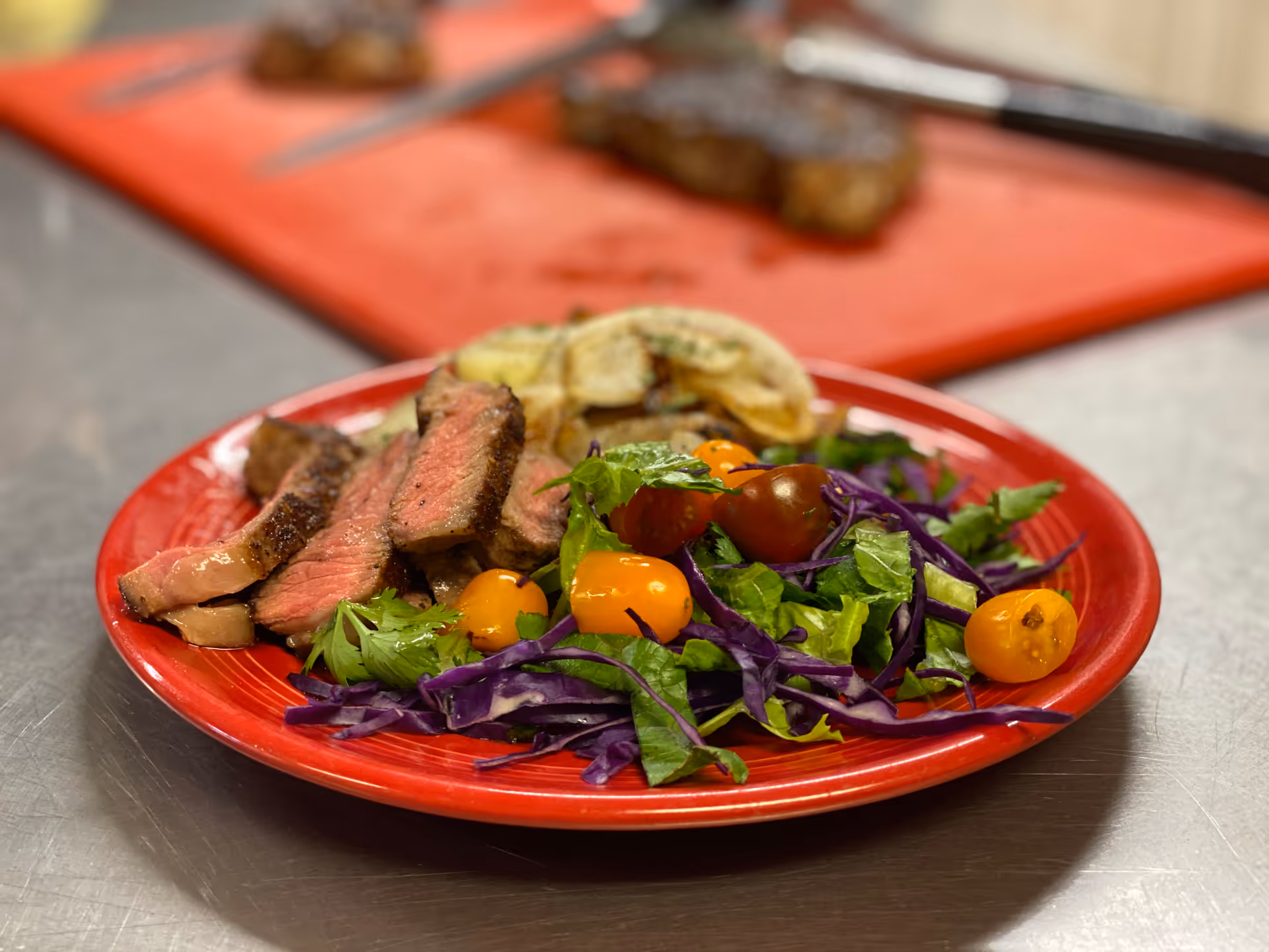 Red plate with sliced steak and colorful salad on stainless counter.