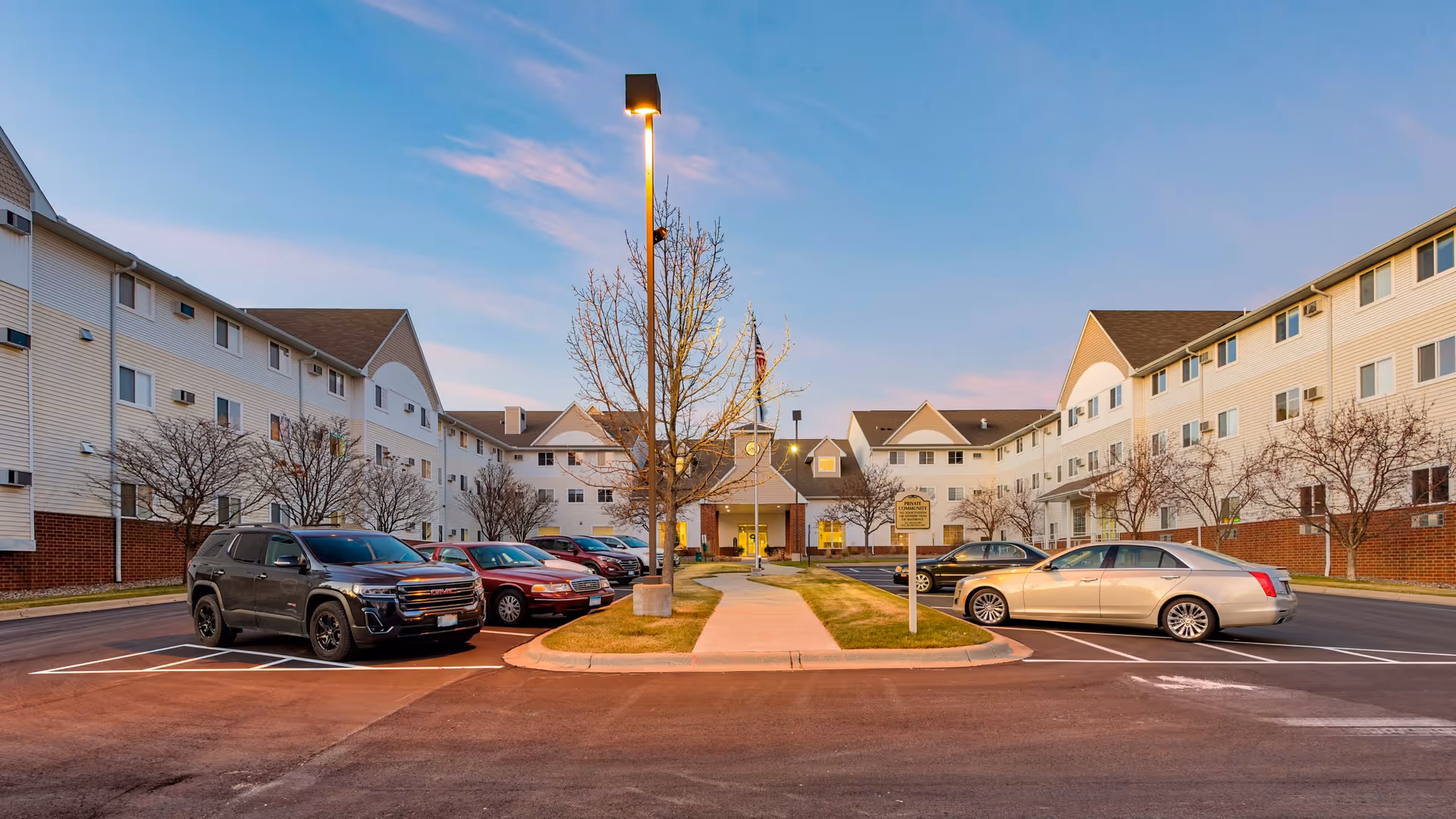 Front driveway and main entrance of a three-story senior living building with parked cars and a central walkway at dusk.