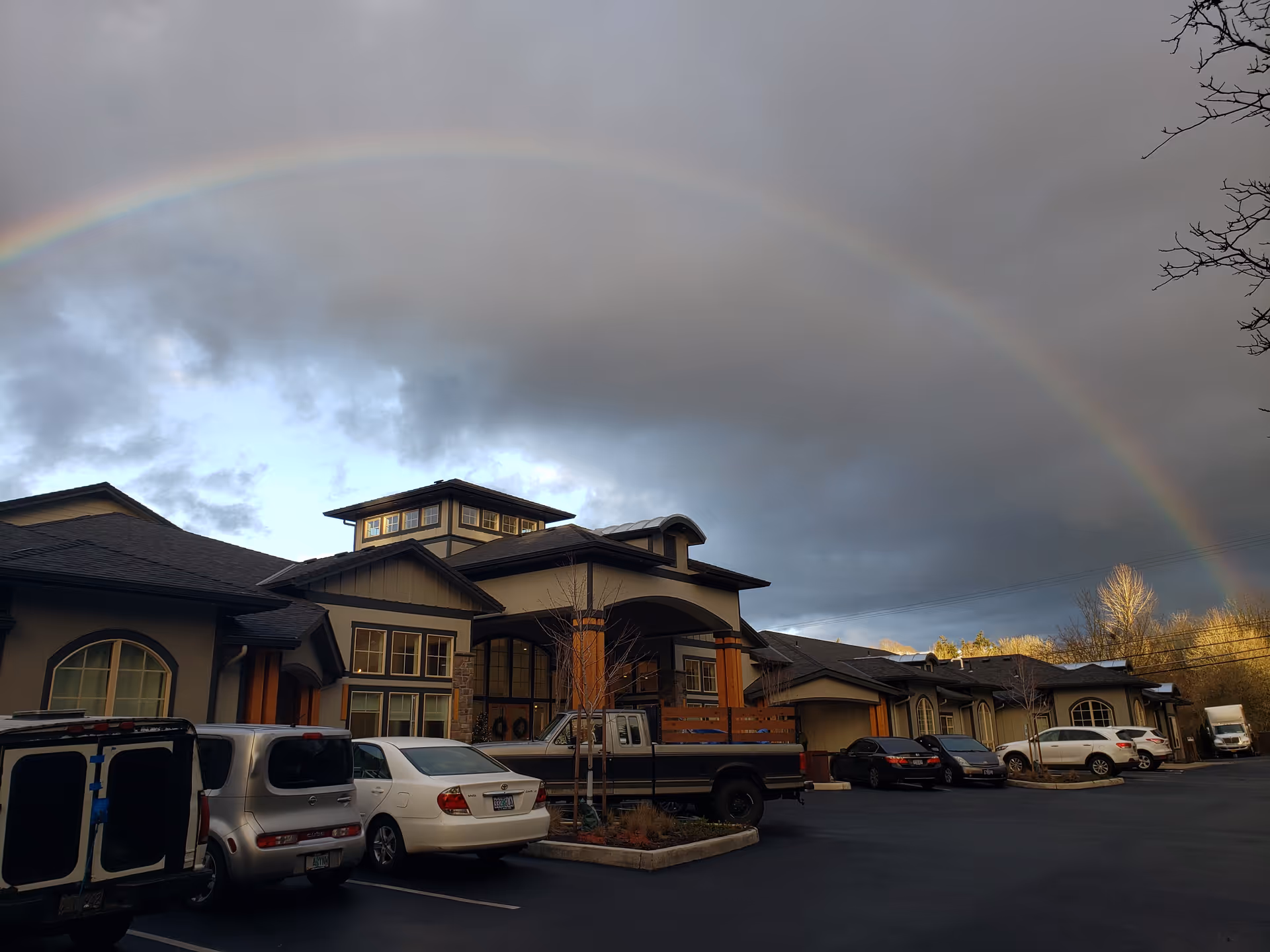 Brookside Memory Care exterior with parked cars and a rainbow across the cloudy sky.