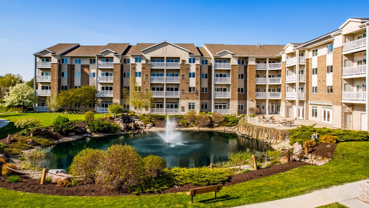 Exterior view of Coventry Village, a multi-story senior living facility with balconies. In front of the building is a landscaped garden area featuring a pond with a water fountain, surrounded by green grass, bushes, and trees. There is a bench and a paved walkway near the pond.