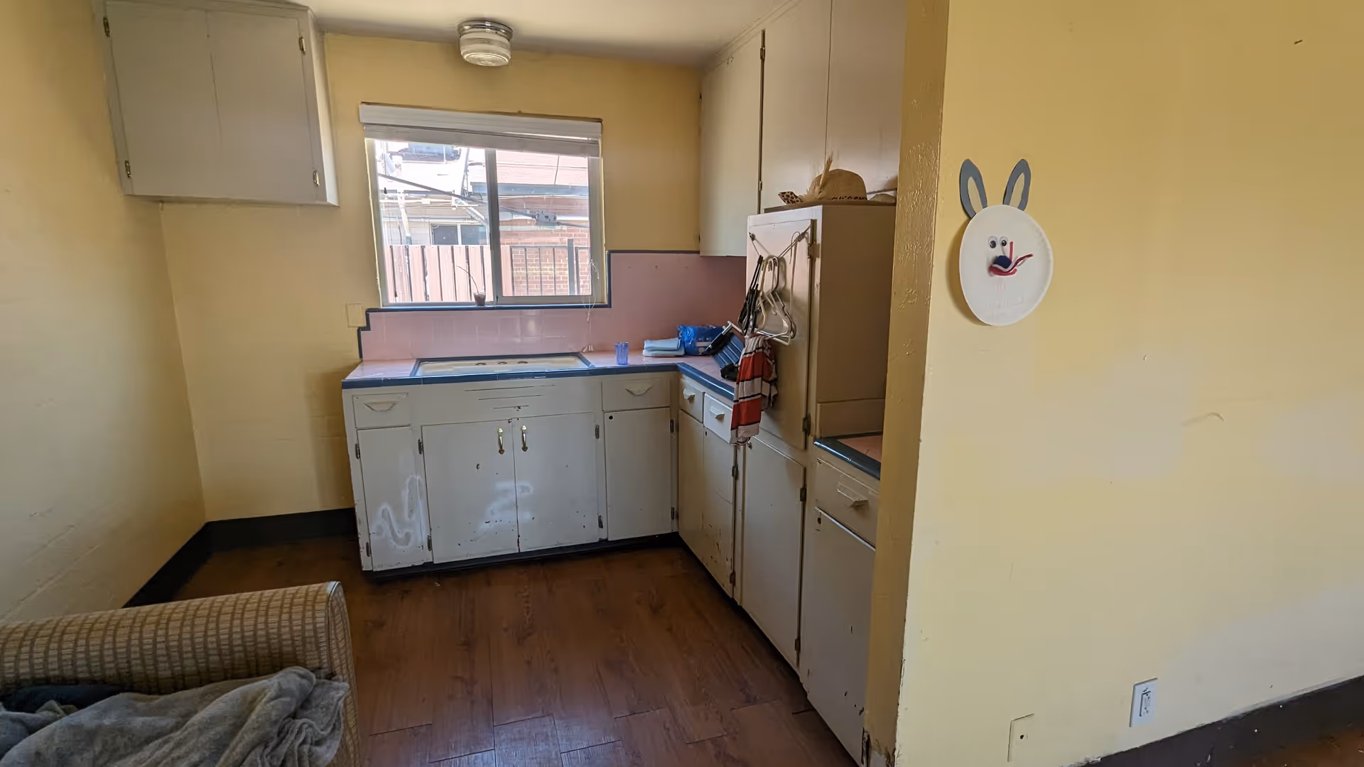 Small, dated kitchen with white cabinets, pink backsplash, and a window view.