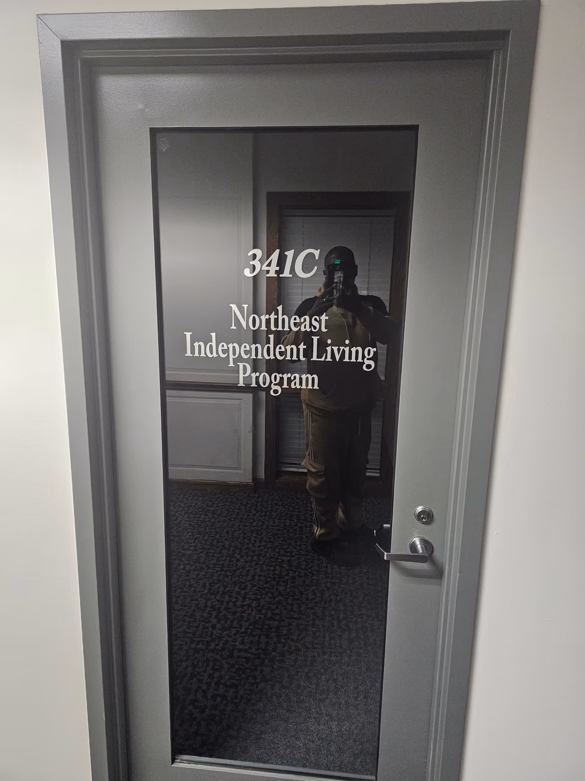 Glass door with white text 341C Northeast Independent Living Program reflecting a person taking a photo