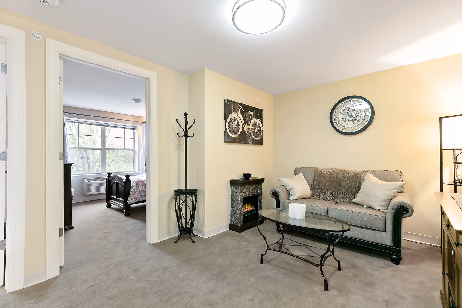 A cozy living room area with a gray sofa adorned with two light-colored pillows and a throw blanket. In front of the sofa is a glass coffee table with three white candles. On the wall above the sofa is a large round clock. To the left of the sofa is a small electric fireplace with a decorative bowl on top. A black metal coat rack stands next to the fireplace. The room has beige walls and carpeted floors. An adjacent bedroom with a large window and a dark wooden bed frame is visible through an open doorway.