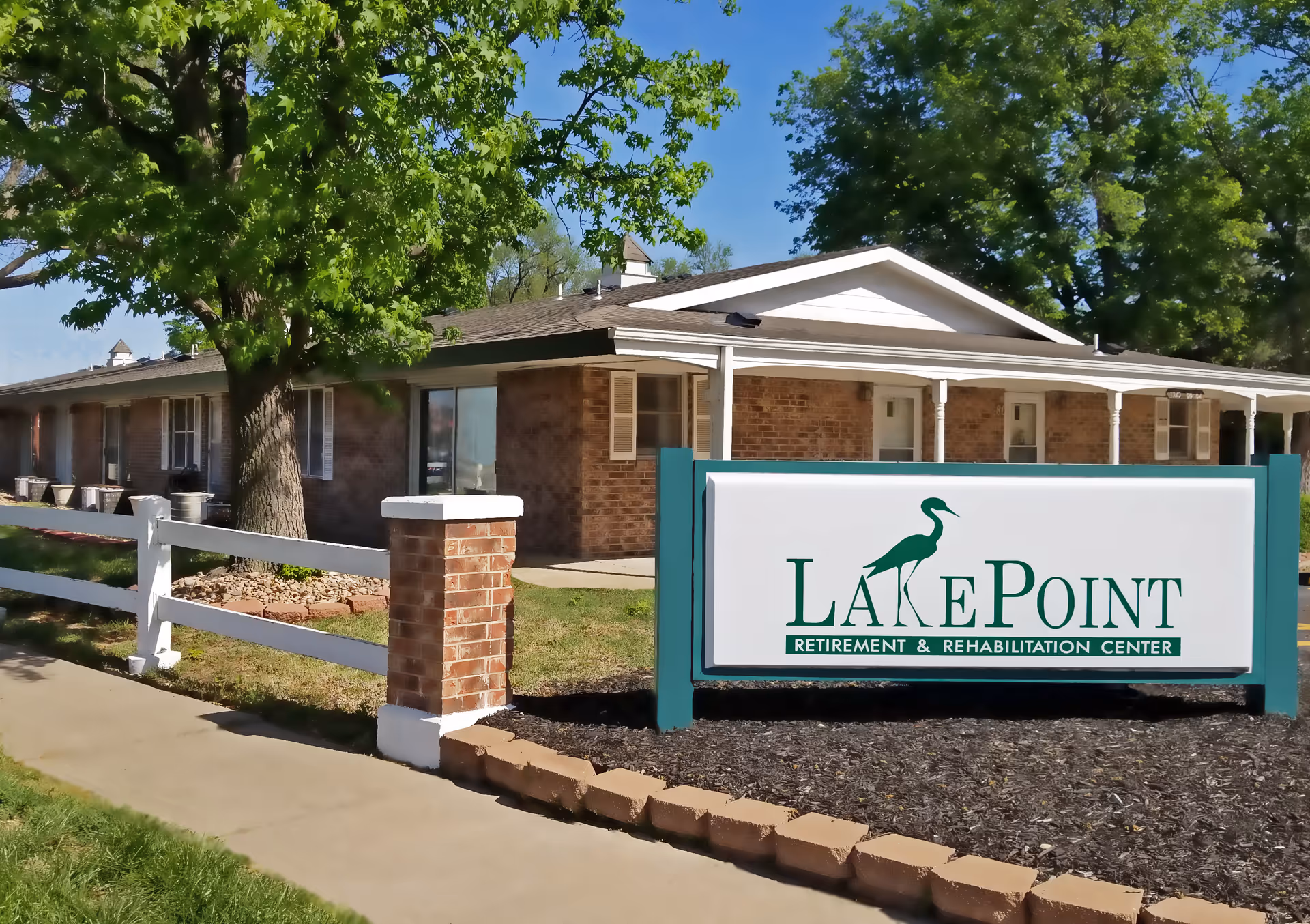 Exterior view of LakePoint Retirement & Rehabilitation Center, a single-story brick building with a white fence, green trees, and a clear blue sky. A large sign with the facility's name and logo is prominently displayed in front.