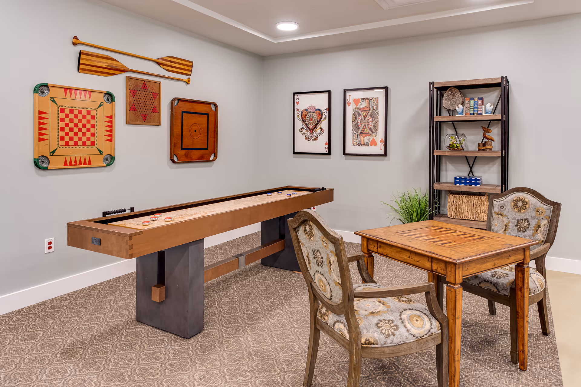 Cozy interior game room with a shuffleboard table, a wooden card table and upholstered chairs, wall-mounted games and shelving.
