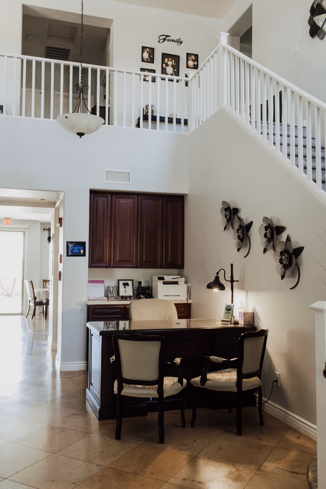 Reception desk with chairs, dark wood cabinetry, and a staircase in a bright assisted living interior.
