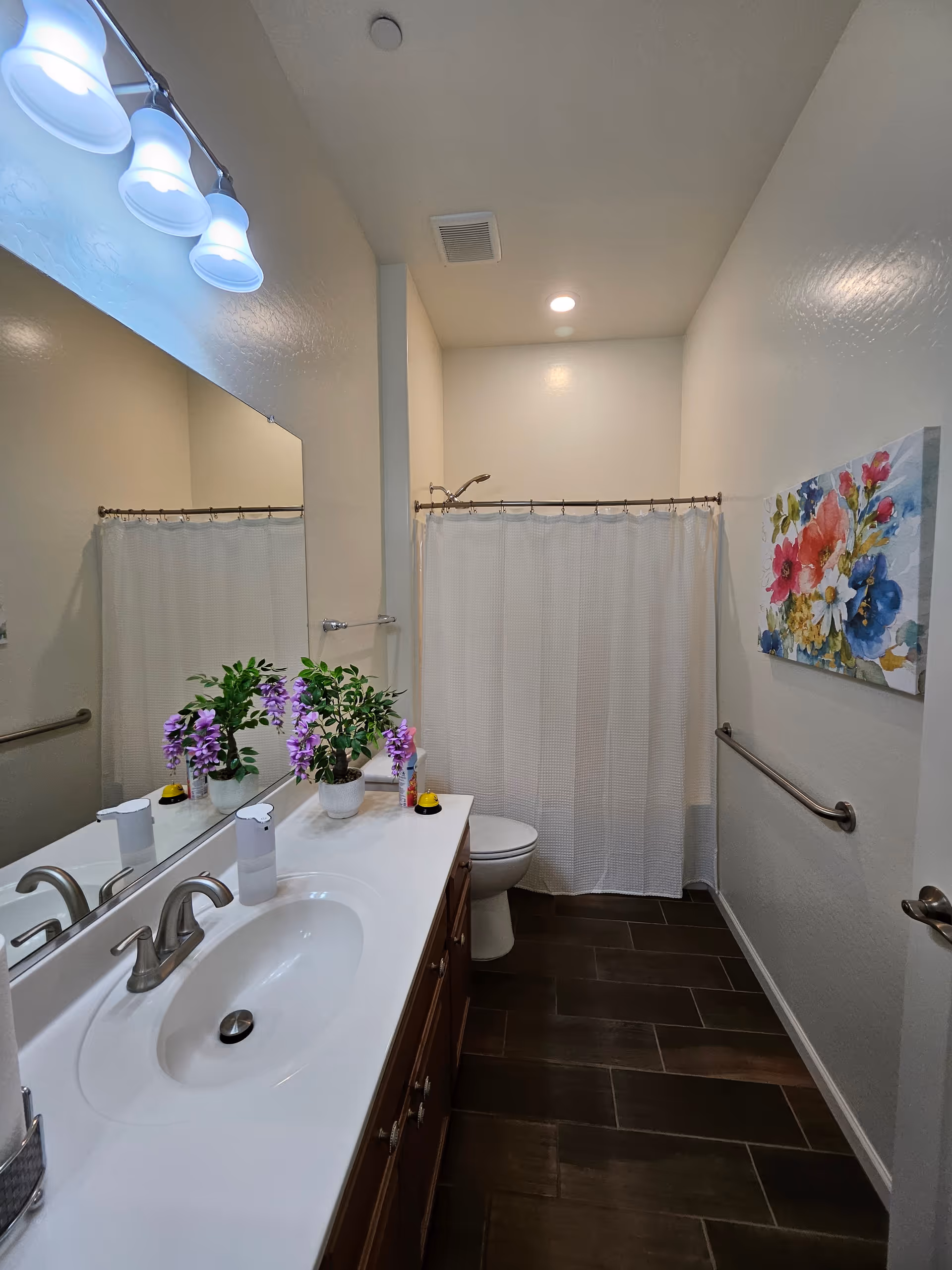 Accessible bathroom with double sink, mirror, flowers, shower curtain, and safety rails.