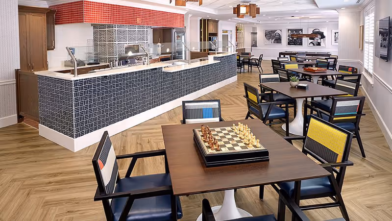 Bright communal dining and activity room with multiple tables and chairs, a central serving counter, and a chessboard on the nearest table.