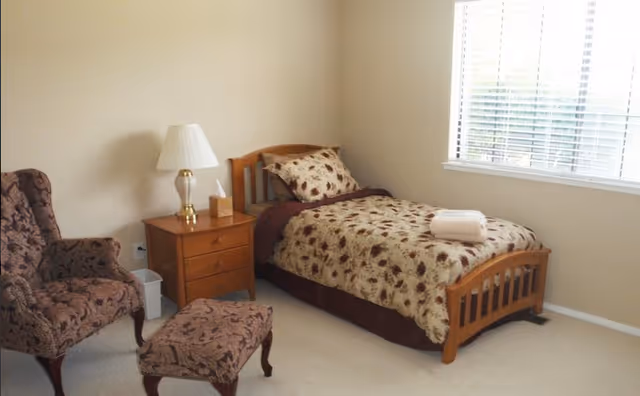 A small bedroom with a single wooden bed covered with a floral patterned bedspread and matching pillow. A rolled white towel is placed on the bed. Next to the bed is a wooden nightstand with a lamp and a tissue box. There is a patterned armchair with a matching footstool near the nightstand. A window with blinds is on the right wall, letting in natural light.