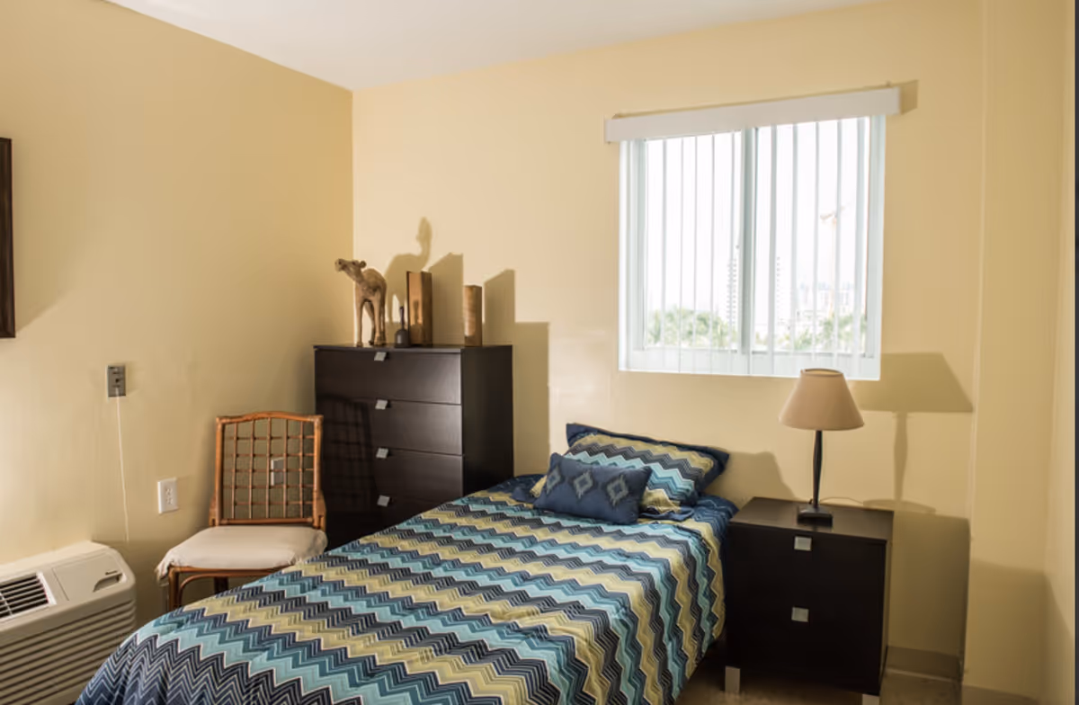 Small bedroom with a single bed in blue zigzag bedding, a nightstand with a lamp, dresser, chair, and a window with vertical blinds.