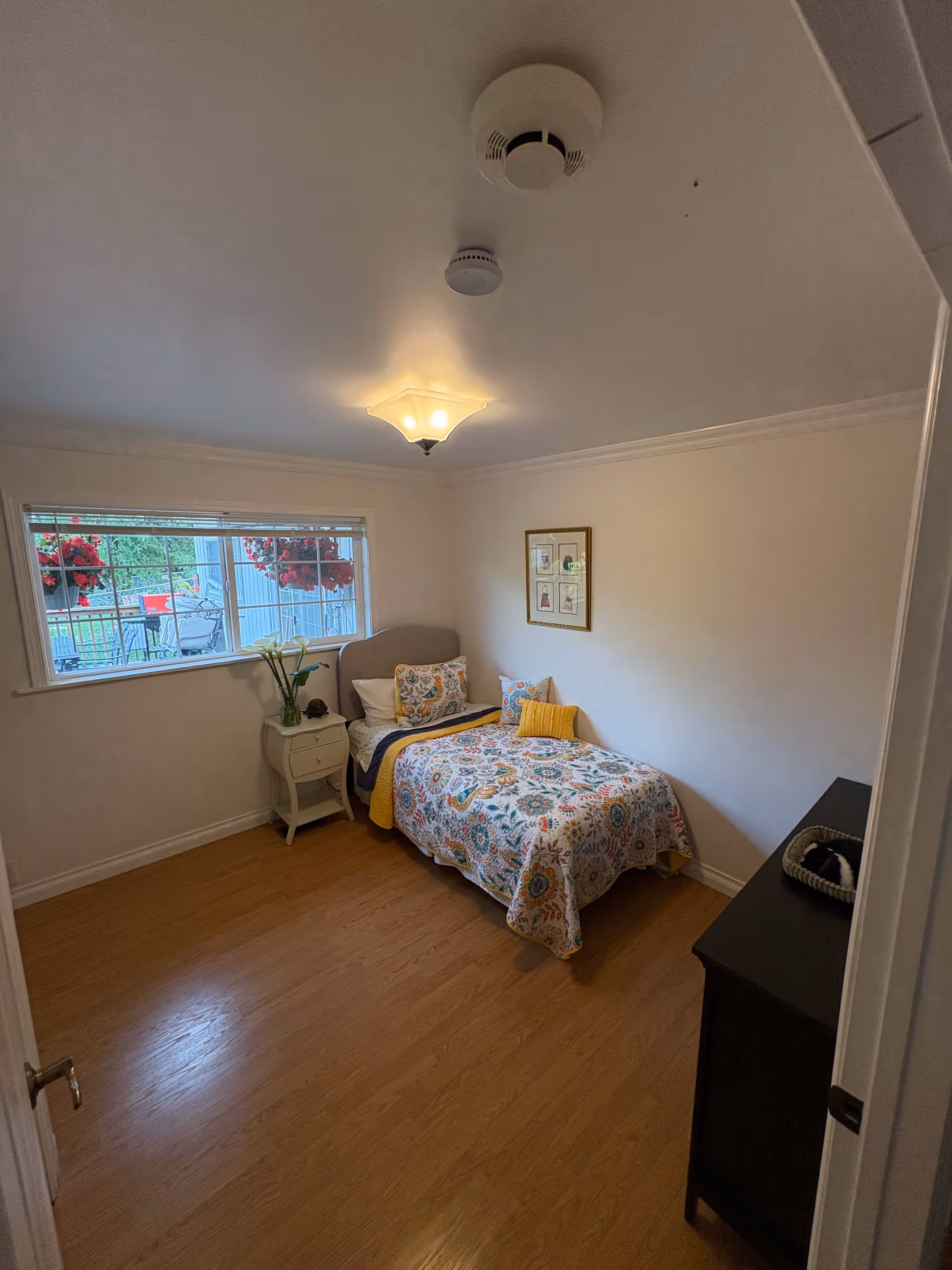 A small, cozy bedroom with a single bed covered in a colorful floral quilt and several pillows. There is a small white nightstand with a plant and a decorative item next to the bed. A window with a view of hanging red flowers and outdoor furniture lets in natural light. The room has light-colored walls, wooden flooring, a ceiling light fixture, and a smoke detector on the ceiling. A dark dresser is partially visible on the right side of the image.