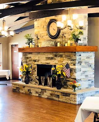 Interior view of a room with a large stone fireplace decorated with a wooden mantel. On the mantel, there are plants, a clock, and a welcome sign. The fireplace hearth has two vases with yellow flowers and small American flags, along with decorative letters spelling 'SVM'. The room has wooden floors, exposed ceiling beams, and chandeliers with multiple lights. Tables with white tablecloths are partially visible.