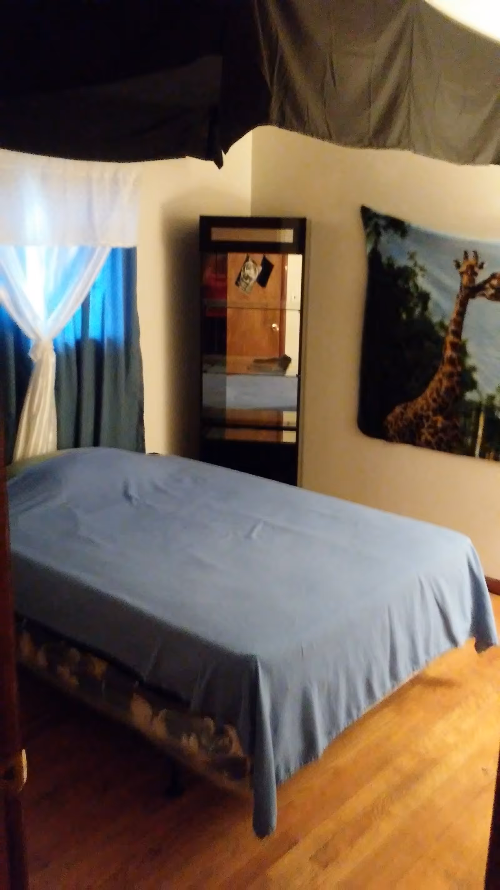 A bedroom with a bed covered in a blue sheet, a window with white and blue curtains, a tall mirror reflecting part of the room, and a wall hanging featuring a giraffe in a natural setting.