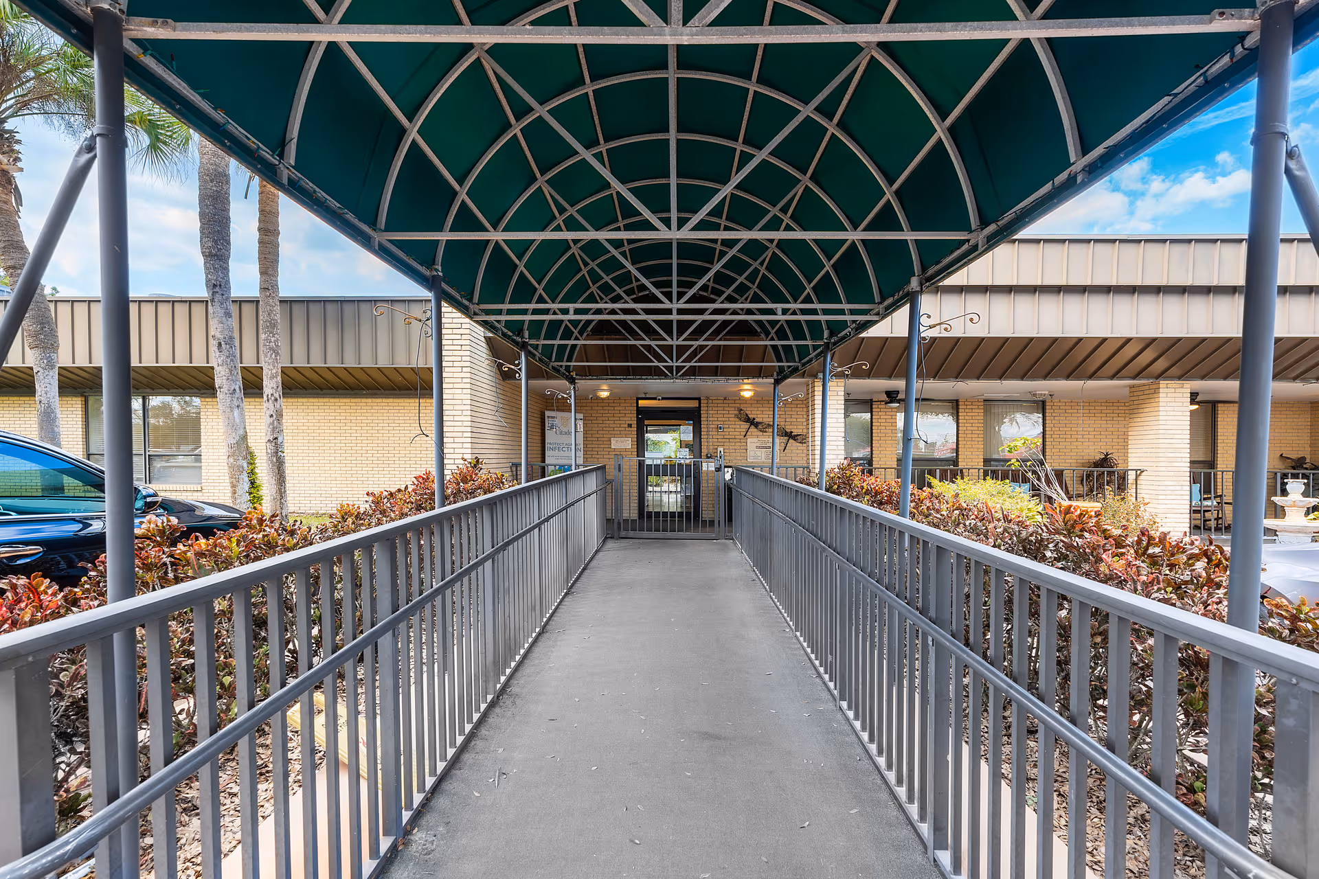 Covered metal-and-fabric walkway ramp leading to the entrance of a brick nursing facility with landscaping on either side.