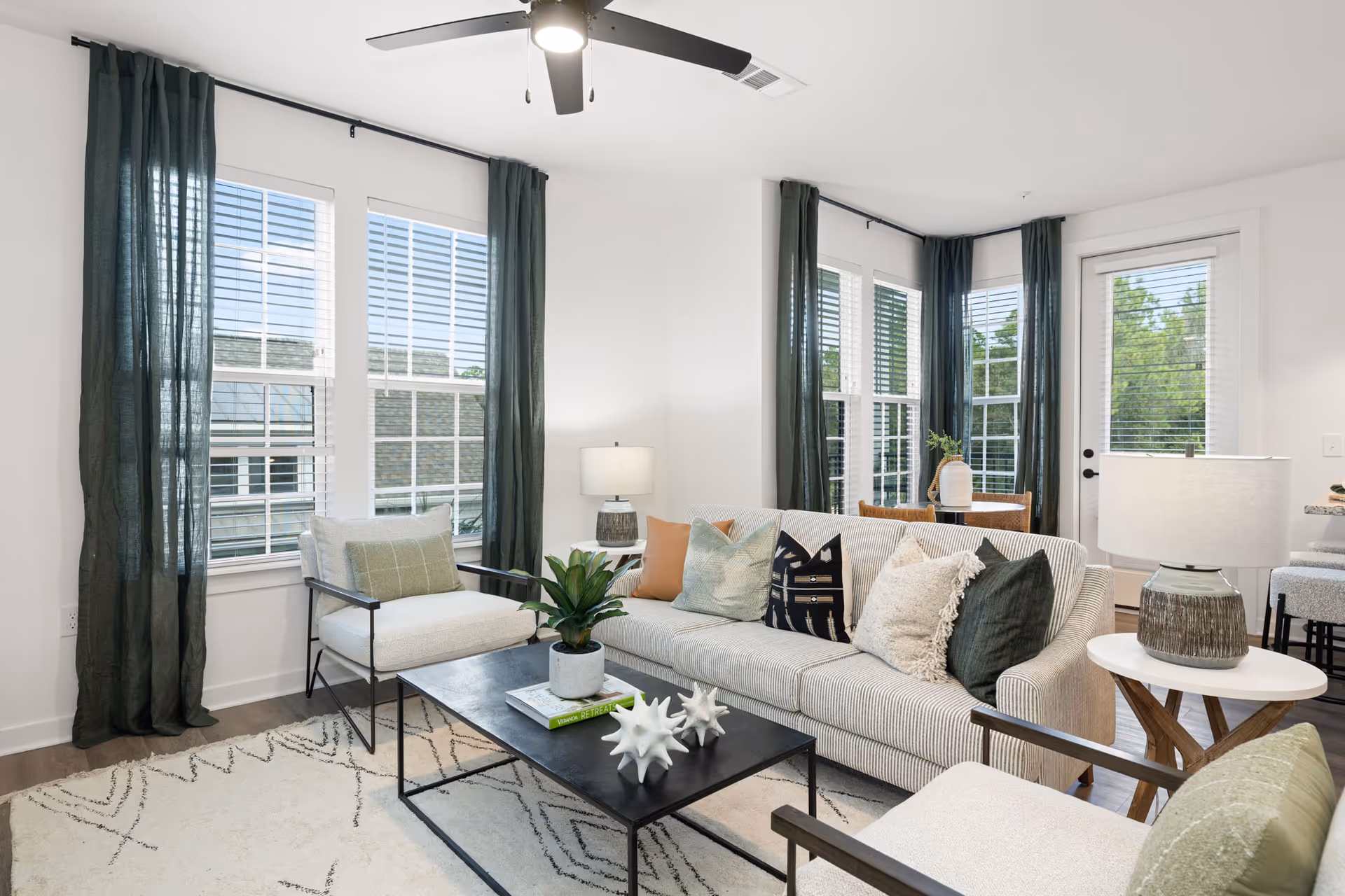 Bright and modern living room with a light-colored sofa adorned with multiple decorative pillows, two matching armchairs with green cushions, a black metal coffee table with decorative items and a plant, large windows with dark green curtains, a ceiling fan, and two side tables with lamps.