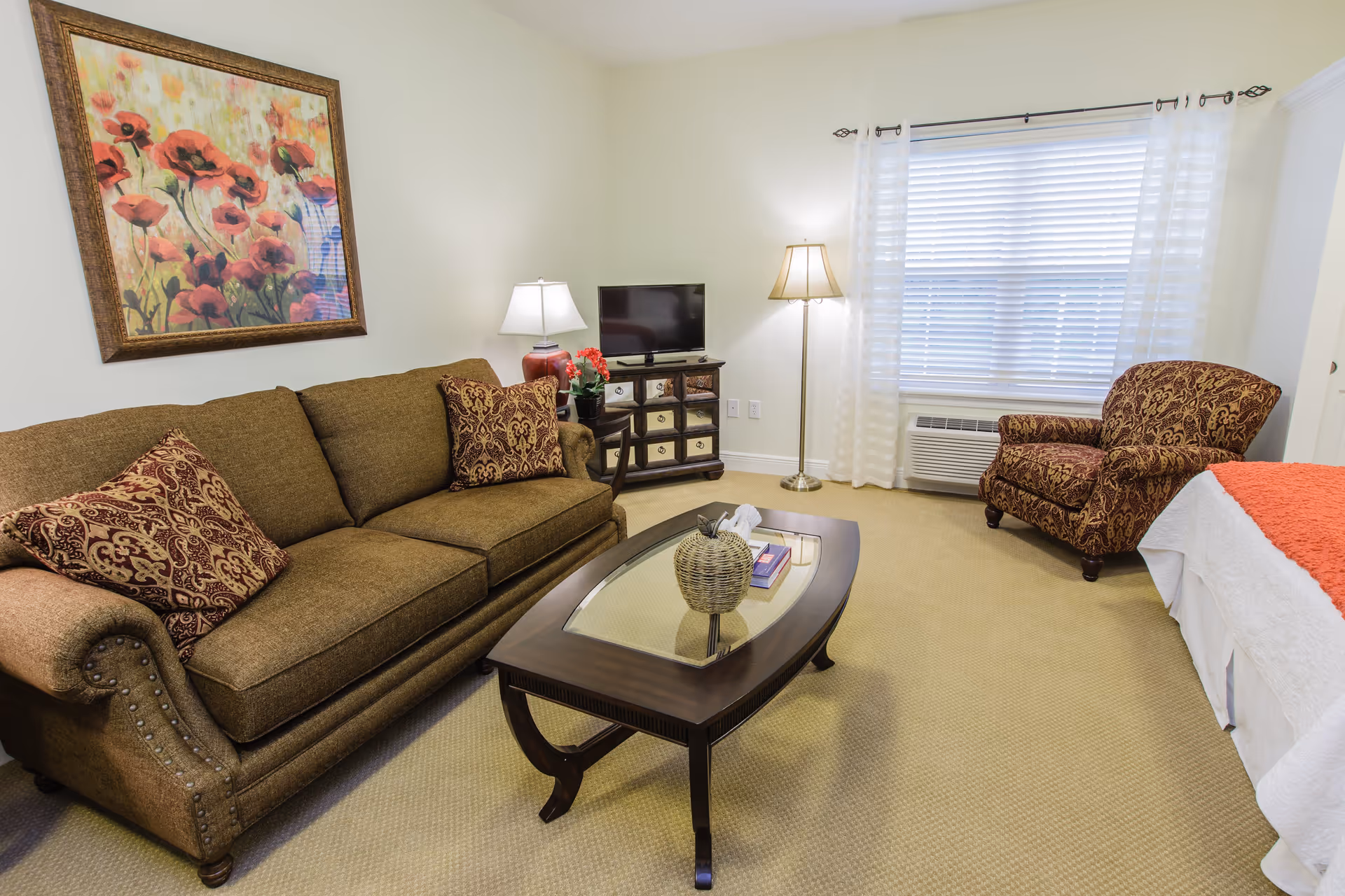A cozy living area in a senior living facility featuring a brown sofa with patterned cushions, a matching armchair, a wooden coffee table with decorative items, a TV on a small cabinet, a floor lamp, and a window with white blinds and sheer curtains. A colorful floral painting hangs on the wall above the sofa.