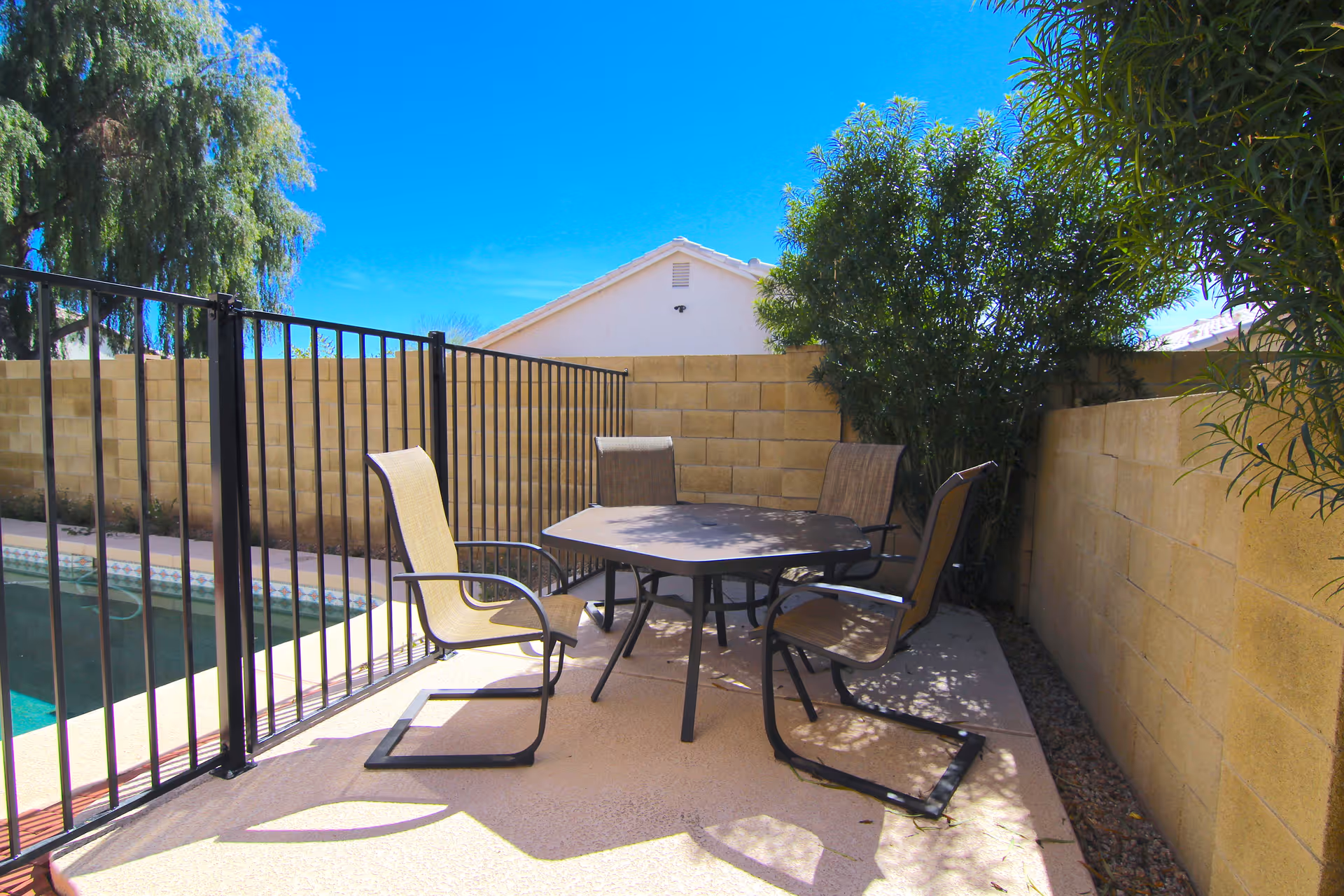 Patio seating area with a round table and four chairs beside a fenced swimming pool and a cinderblock wall.