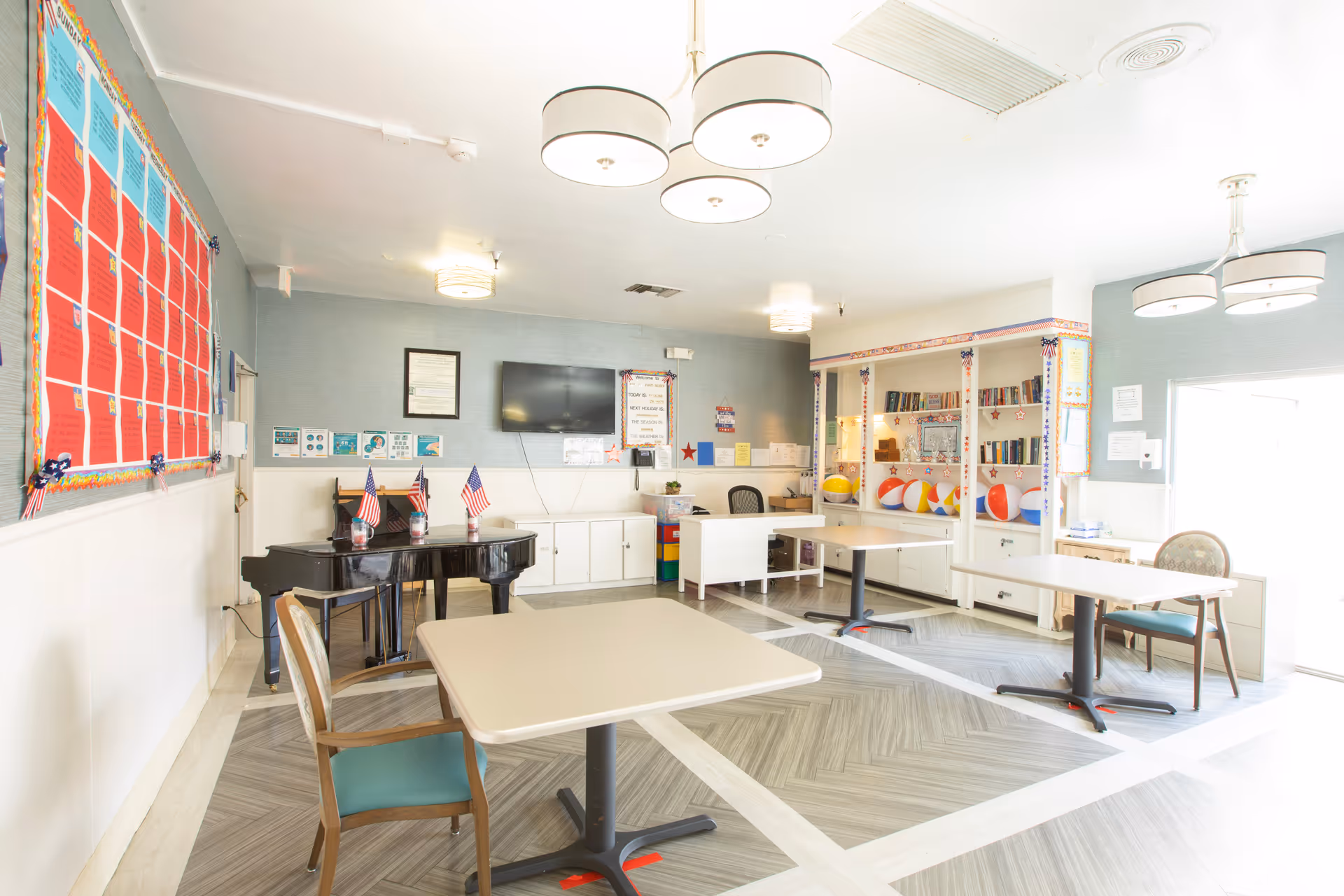 Bright senior activity/common room with tables and chairs, a piano with small American flags, a TV, bulletin board and shelving with books and beach balls.