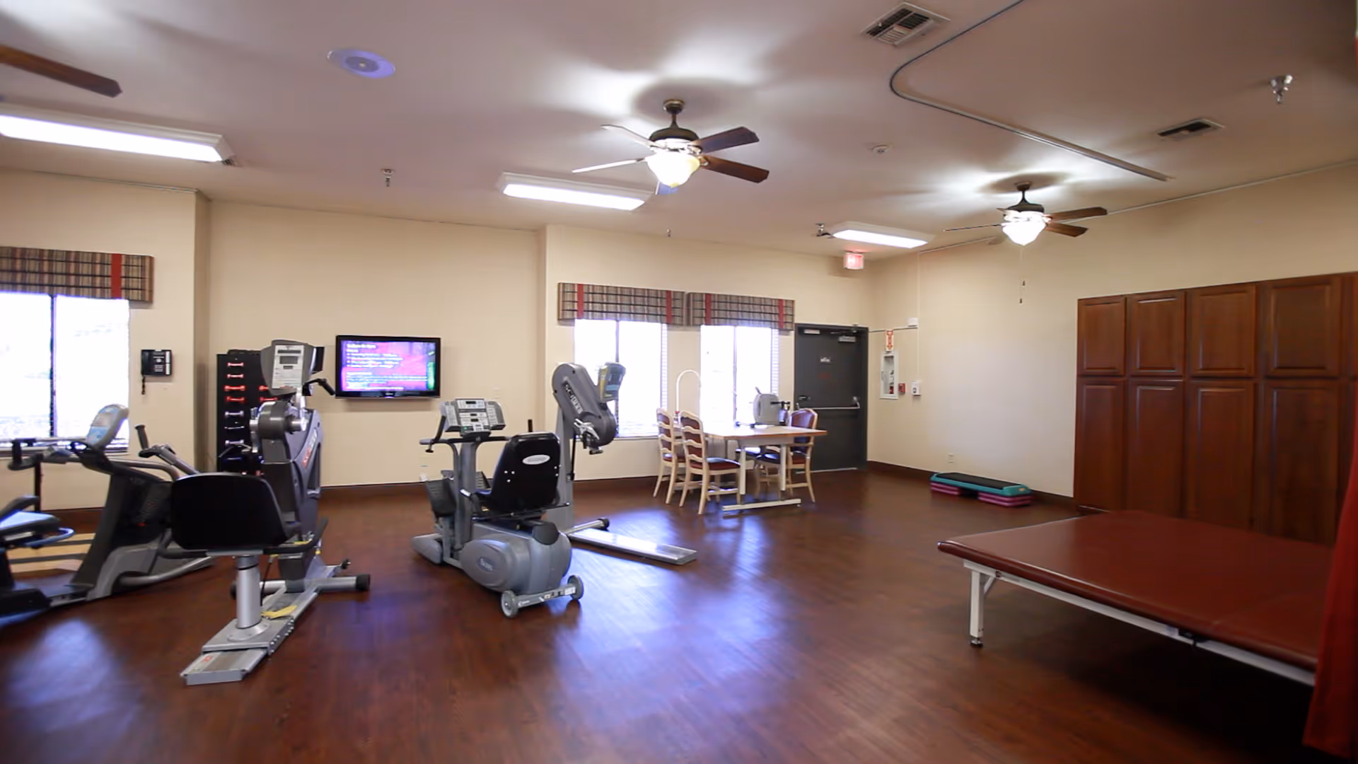 A spacious exercise room with wooden floors and beige walls featuring several pieces of cardio exercise equipment including stationary bikes and treadmills. There is a wall-mounted TV, a table with chairs near windows with plaid valances, ceiling fans, and a padded therapy table on the right side.