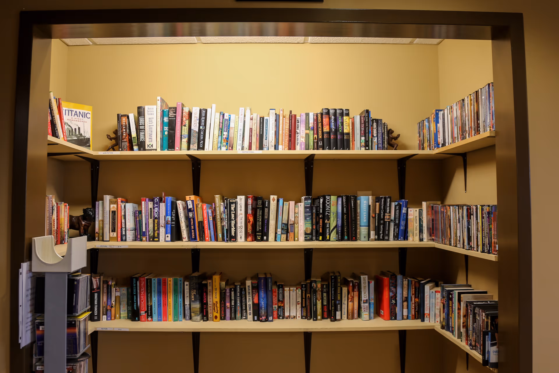A small library or reading nook with three shelves filled with books and DVDs against a beige wall, framed by a dark brown doorway.