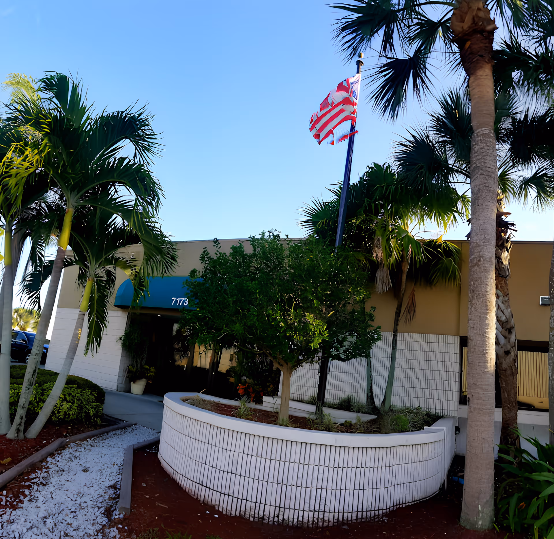 Entrance of a single-story building with palm trees, a raised circular planter, and an American flag flying above.