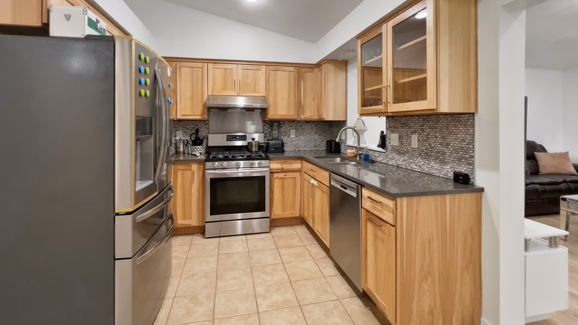 Modern kitchen with wooden cabinets and stainless appliances; living room visible beyond.