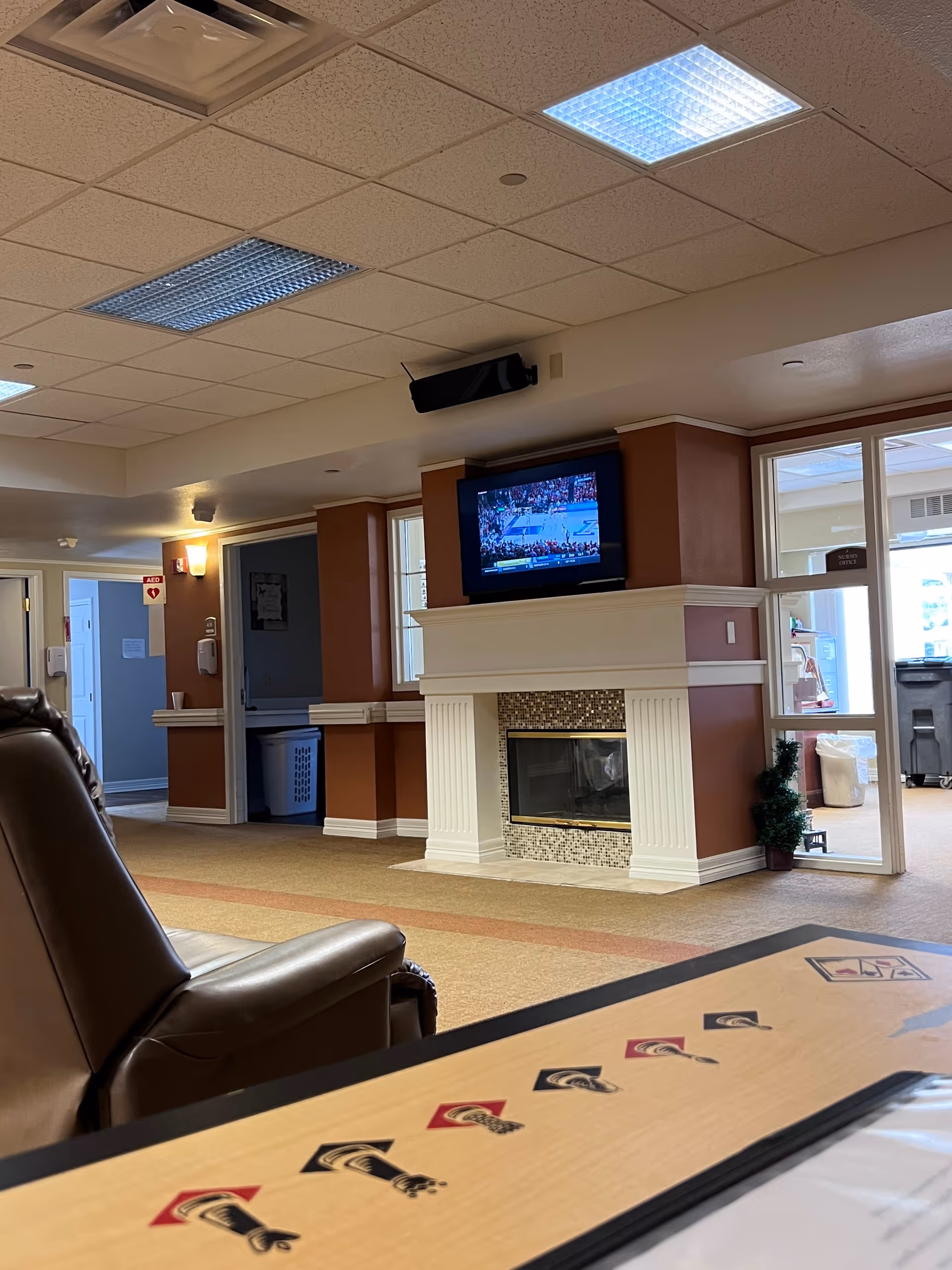 Lounge area with a fireplace, mounted TV, and leather chair in a senior residence.