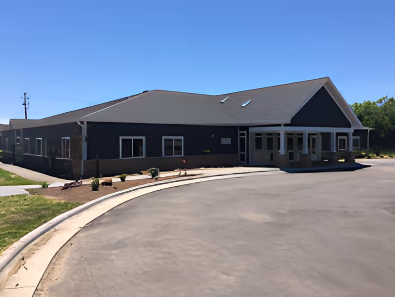 Single-story senior living facility building with a covered main entrance, surrounding driveway, and clear blue sky.