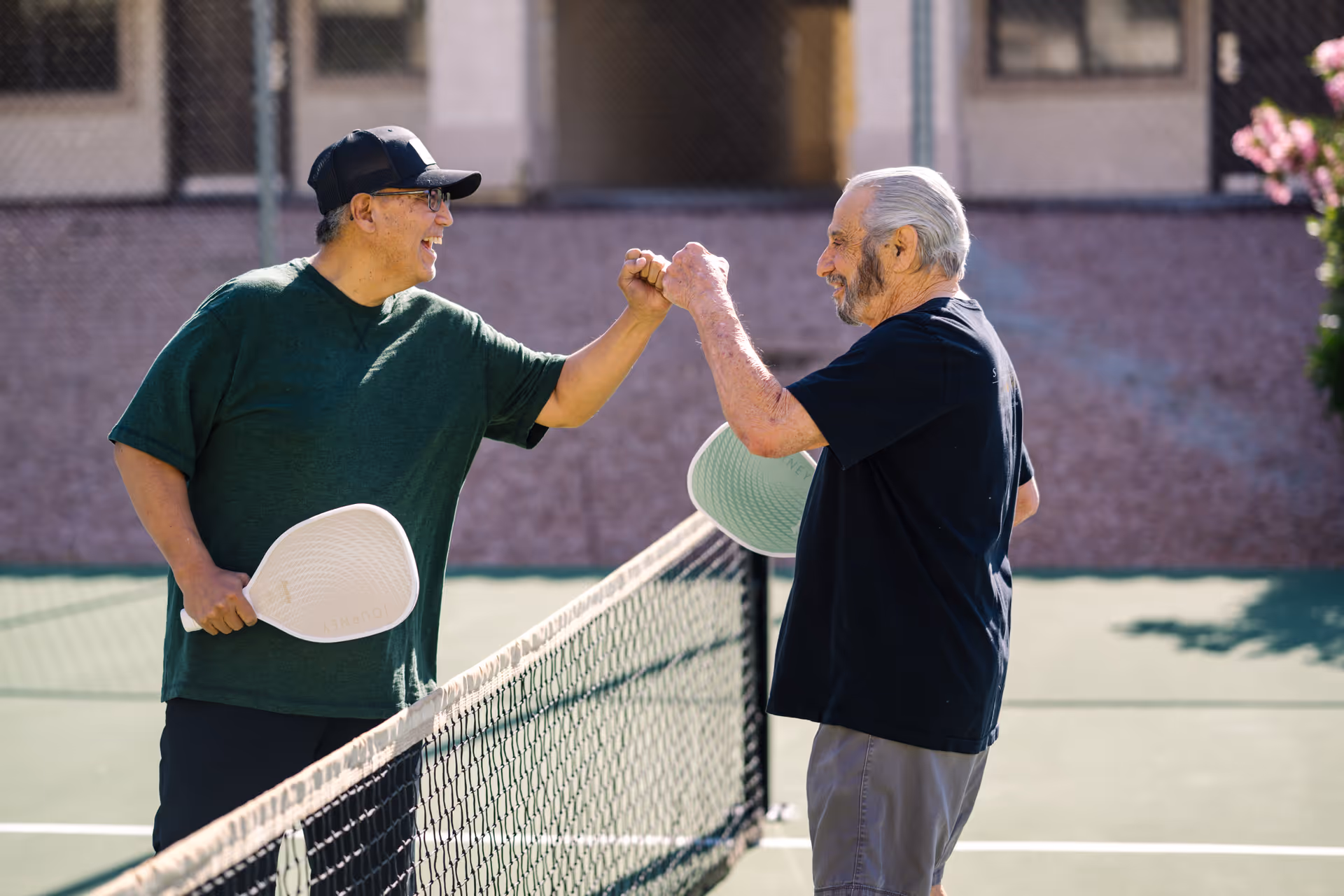 Two elderly men on a tennis court giving each other a fist bump, each holding a pickleball paddle, with a building and chain-link fence in the background.