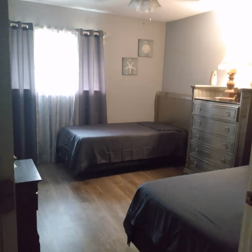 Two twin beds in a gray-toned bedroom with dresser and curtained window.