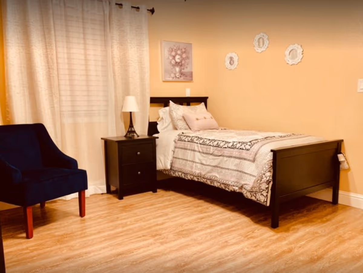 Cozy bedroom with a single bed, black nightstand and lamp, navy armchair, and curtained window.