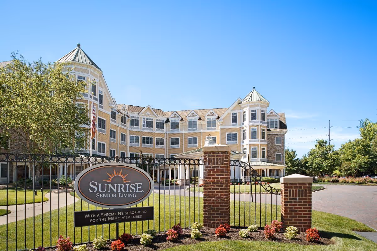 Gated front exterior of the Sunrise Senior Living building with a sign, brick pillars, landscaped flowerbeds, and a clear blue sky.