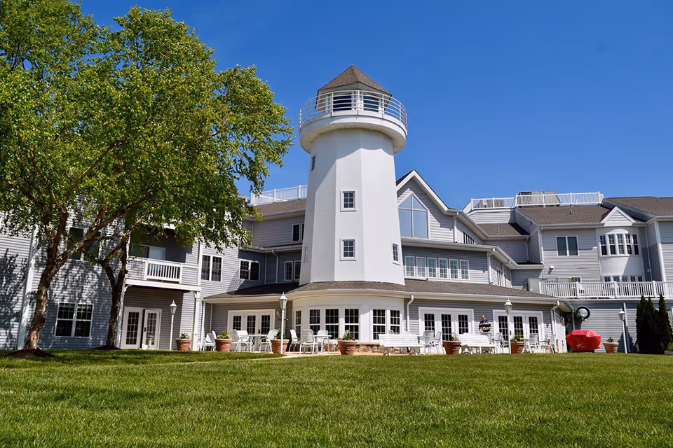 Exterior view of a large assisted living facility with a lighthouse-style tower, multiple windows, outdoor seating with white chairs and tables, potted plants, a large green lawn, and a tree under a clear blue sky.