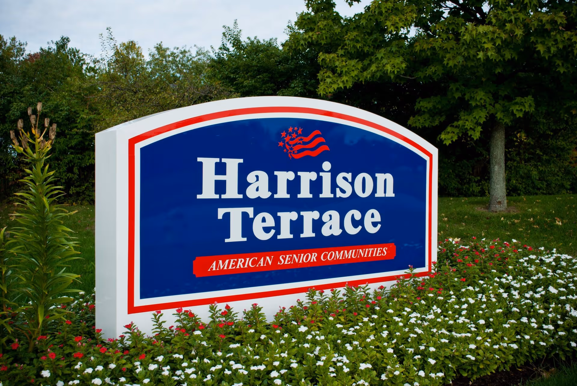 A large outdoor sign for Harrison Terrace, an American senior community, surrounded by green plants and flowers with trees in the background.