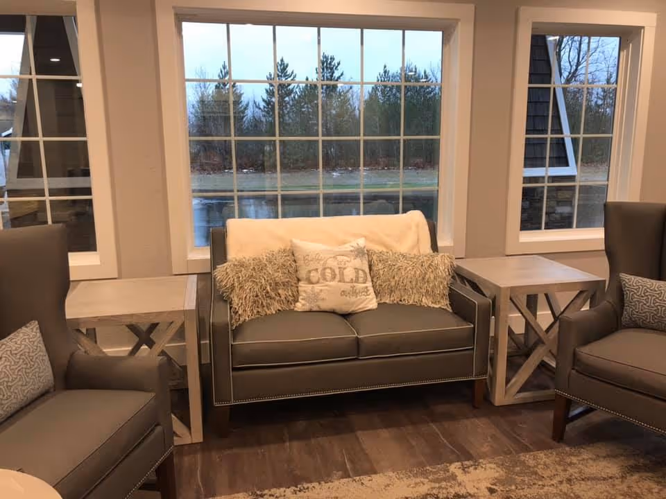 A cozy sitting area with a gray loveseat adorned with three decorative pillows, flanked by two matching gray armchairs each with a patterned cushion. The furniture is arranged around two light-colored wooden side tables. Large windows behind the loveseat reveal an outdoor view of trees and a wet road.