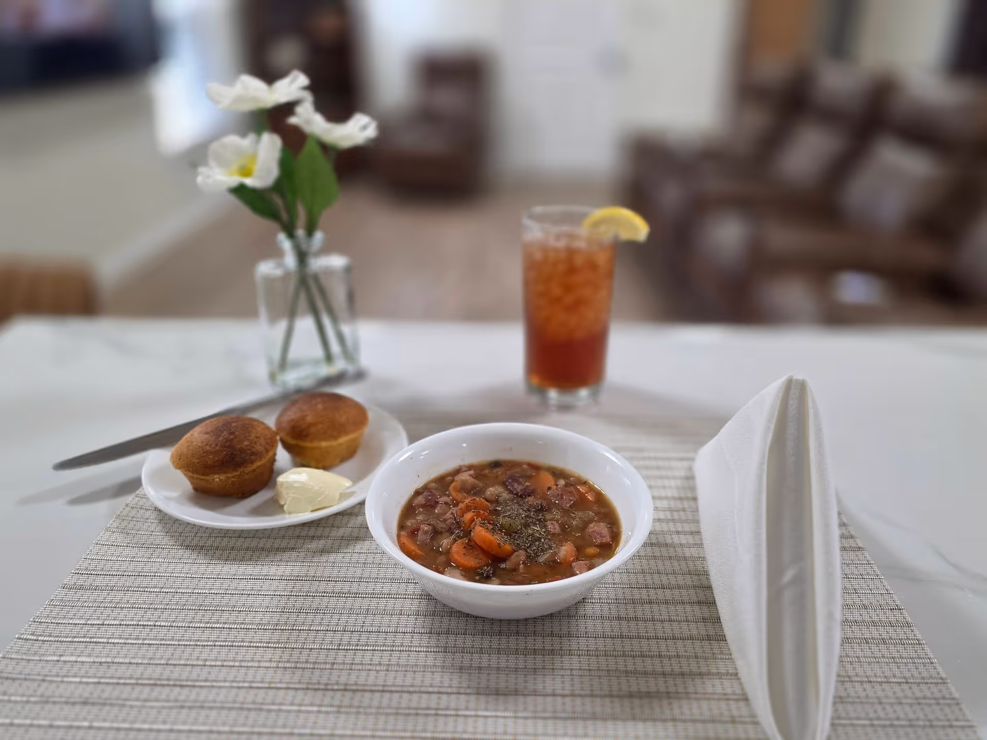 A table set with soup, muffins, drink, napkin, and flowers in a senior living dining room.