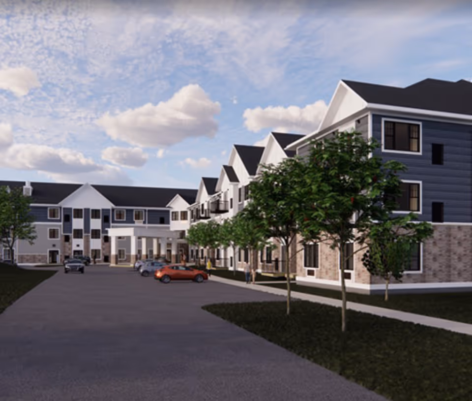 Exterior view of a senior living facility building with multiple floors, a driveway with parked cars, trees along the sidewalk, and a partly cloudy sky.