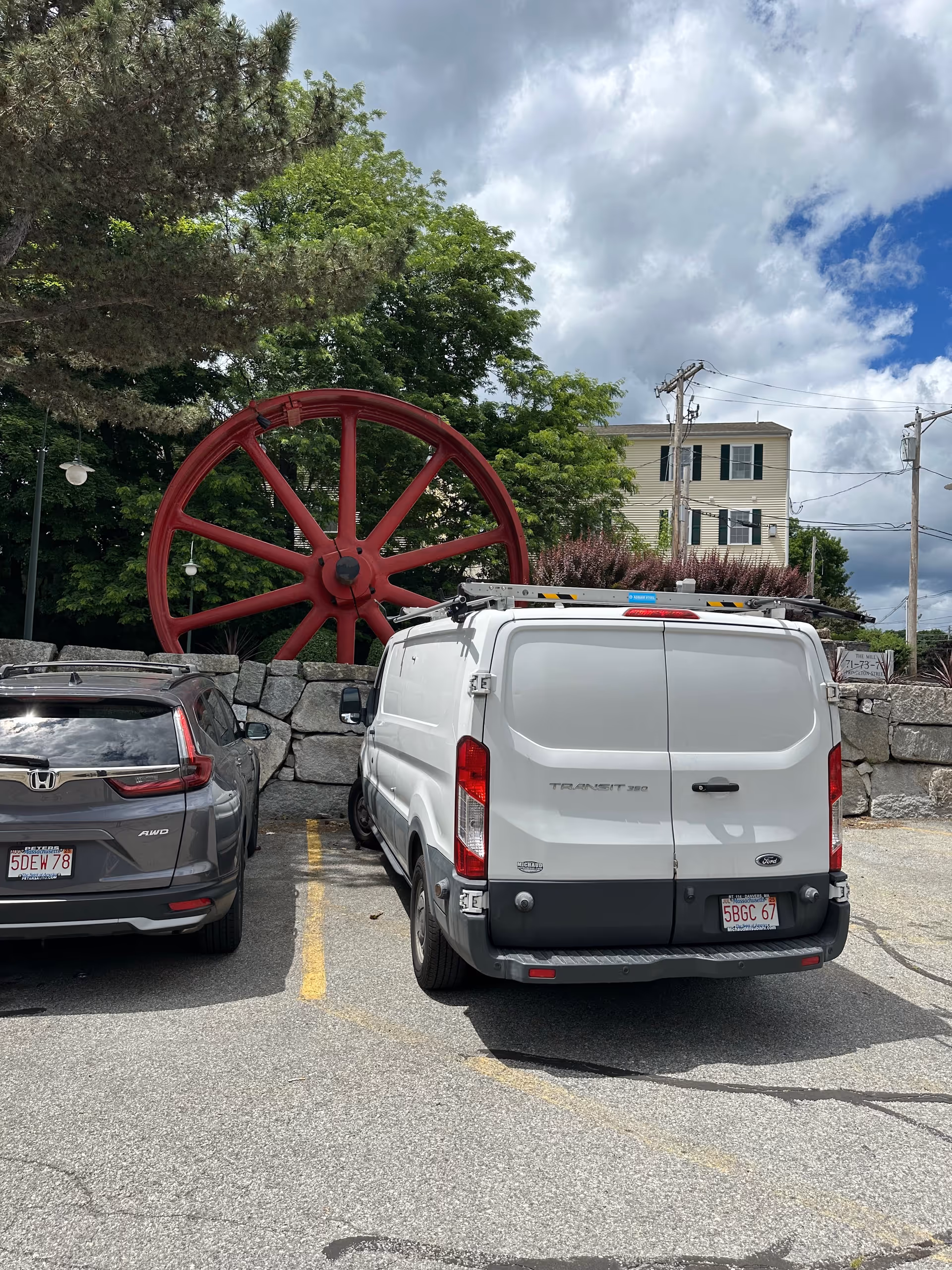 Parking lot with two vehicles and a large red wheel sculpture behind a stone wall.