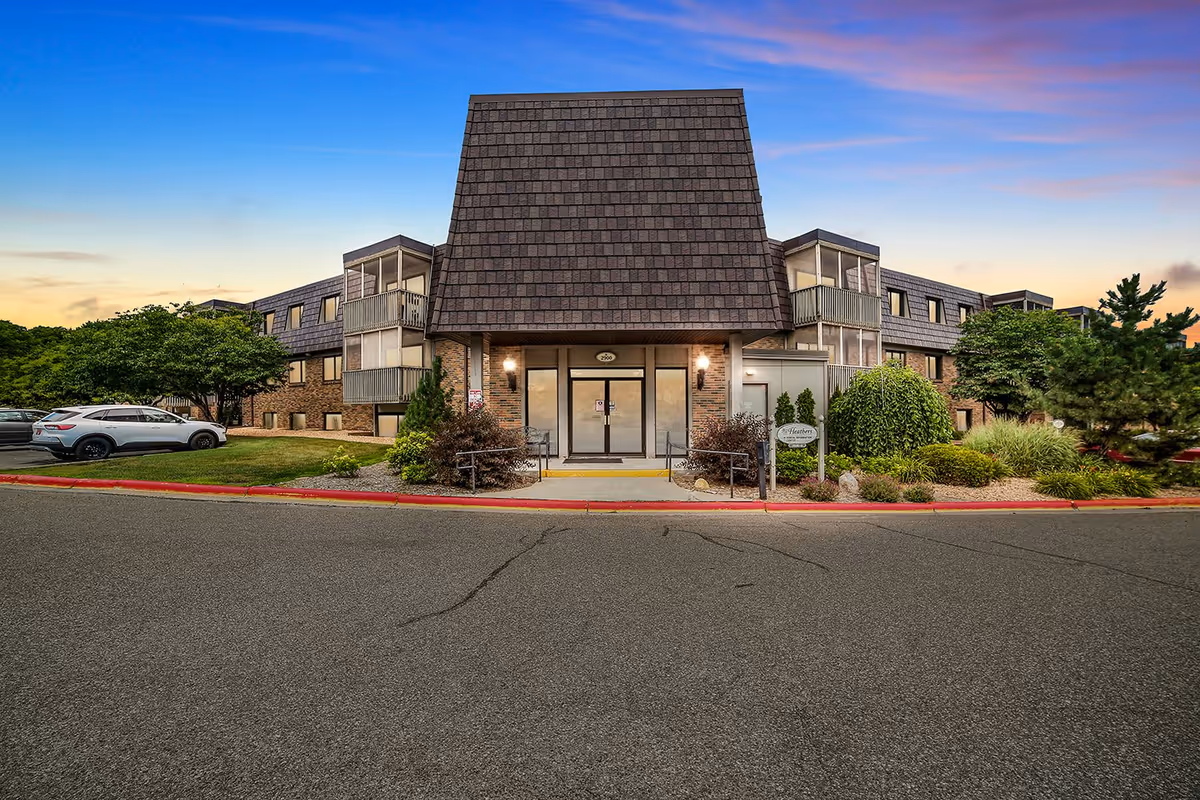 Front exterior view of Heathers Estates building at sunset, showing a three-story structure with a large shingled roof over the entrance, glass doors, surrounding greenery, parked cars, and a clear sky with pink and blue hues.